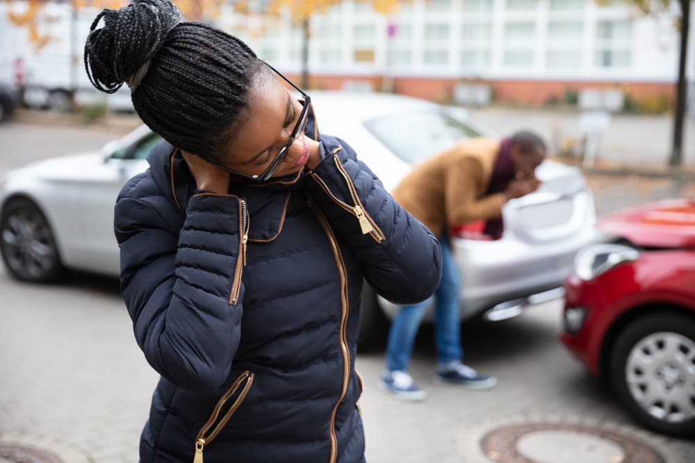 Woman holding her neck in pain while standing near a damaged car, with a man inspecting the vehicle after a road accident.