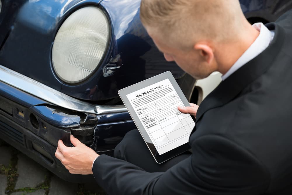An insurance agent inspects a damaged car while reviewing the insurance claim form on a digital tablet.