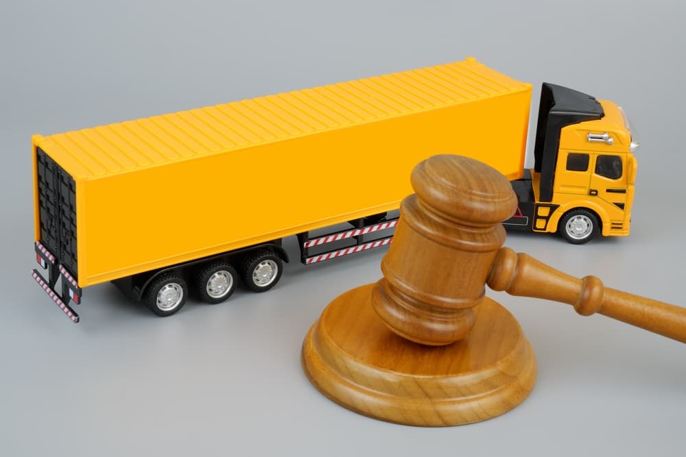 Yellow cargo truck and wooden gavel placed on a grey background, symbolizing legal issues and regulations in the transportation industry.