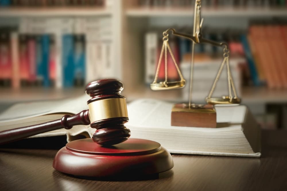 Judge’s gavel and scales of justice on a desk, with a background of a library filled with numerous legal books, symbolizing law and justice.