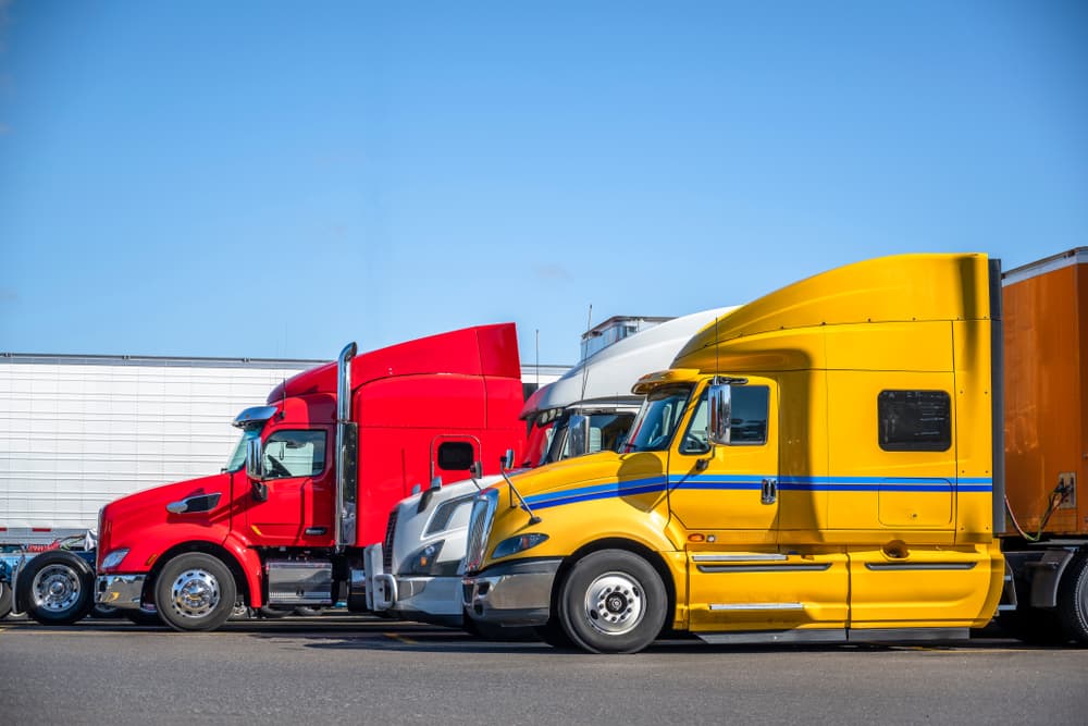 Various makes and colors of big rigs and semi trucks with trailers lined up at a truck stop parking lot, providing a resting space for truck drivers and ensuring compliance with regulations.