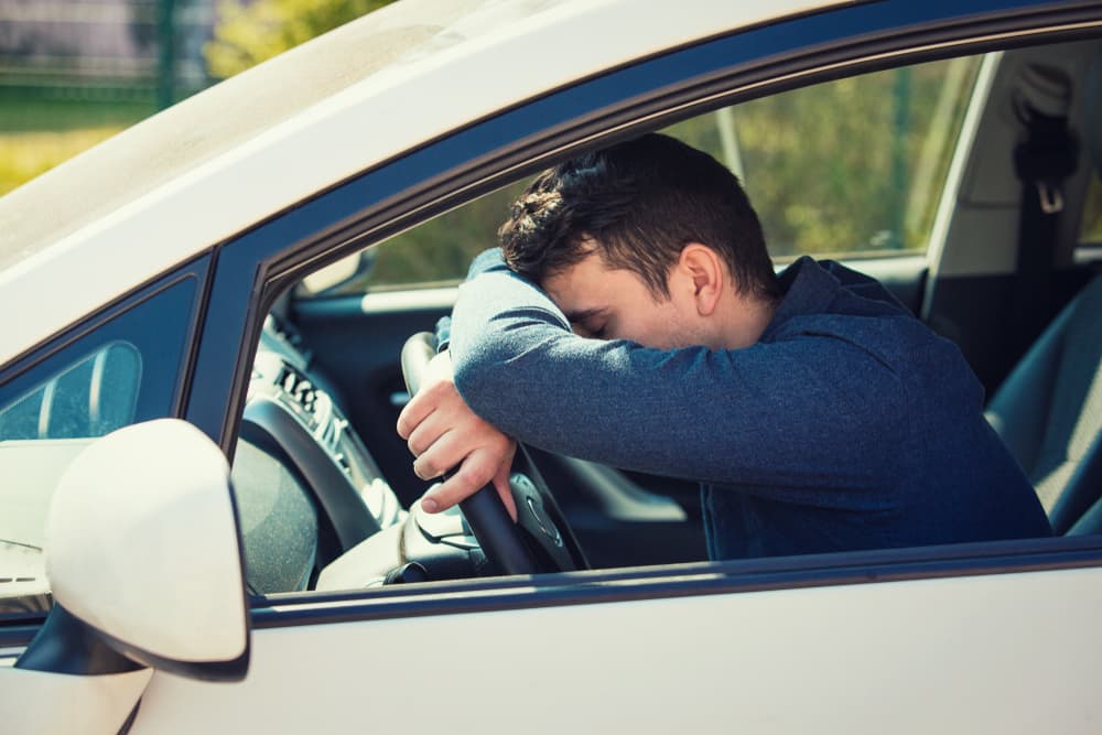 Exhausted young man resting his head and hand on the steering wheel, visibly tired while stuck in rush-hour traffic after work.