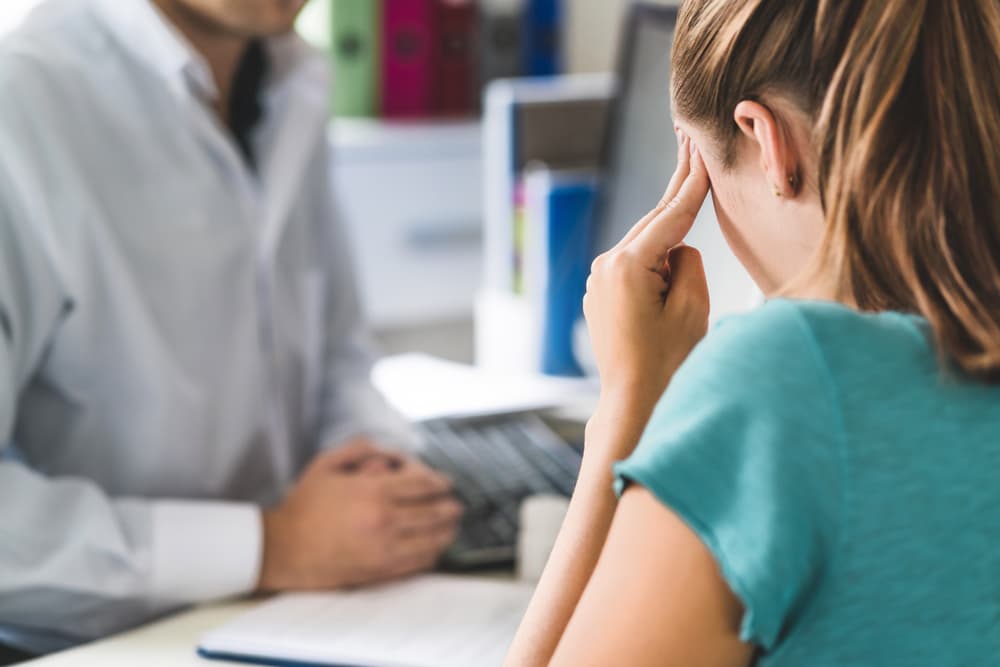 Young woman in pain, holding her head with her hands during a medical appointment, possibly suffering from a migraine, stress, or burnout.