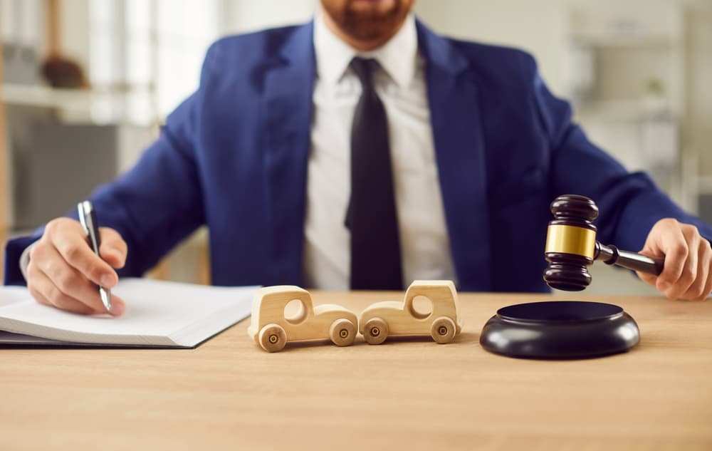 Close-up of wooden toy cars and a judge's gavel with a male lawyer working at a desk in the background, symbolizing car accidents and legal matters.