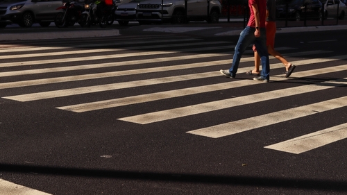 Two pedestrians crossing a city street at a zebra crosswalk during daylight, with cars and motorcycles waiting in the background.